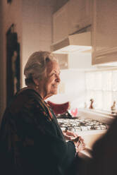 Seitenansicht einer fröhlichen, positiv gestimmten älteren Frau mit grauen Haaren und warmer Kleidung, die am Spülbecken in der Küche steht - ADSF31503