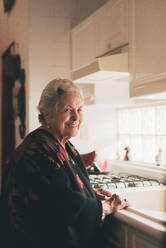 Seitenansicht einer fröhlichen, positiv gestimmten älteren Frau mit grauen Haaren und warmer Kleidung, die am Spülbecken in der Küche steht - ADSF31504
