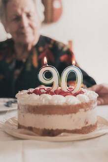 Ältere Frau bläst Kerzen auf Geburtstagskuchen aus und klatscht in die Hände, während sie mit Verwandten zu Hause ihren 90. Geburtstag feiert - ADSF31507