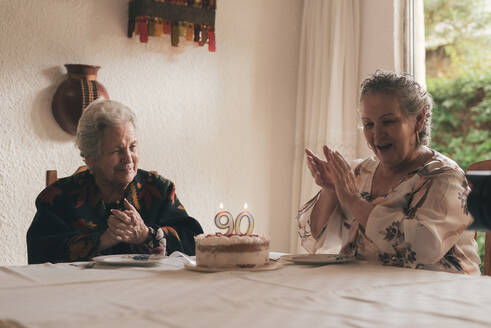 Ältere Frau mit grauen Haaren und ältere Frau sitzen am Esstisch und feiern ihren 90. Geburtstag mit leckerem Kuchen mit Kerzen - ADSF31508
