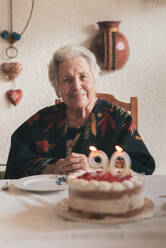 Aged woman blowing candles on birthday cake then clapping hands while celebrating 90th anniversary with relative at home - ADSF31509