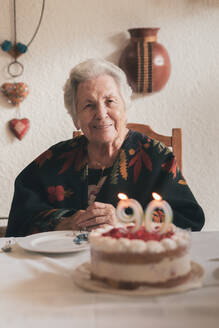 Ältere Frau bläst Kerzen auf Geburtstagskuchen aus und klatscht in die Hände, während sie mit Verwandten zu Hause ihren 90. Geburtstag feiert - ADSF31509