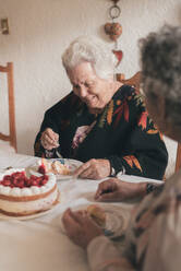 Elderly woman with gray hair and senior female sitting at dining table and celebrating 90th birthday with delicious cake with candles - ADSF31510