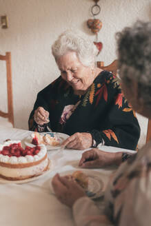 Ältere Frau mit grauen Haaren und ältere Frau sitzen am Esstisch und feiern ihren 90. Geburtstag mit leckerem Kuchen mit Kerzen - ADSF31510