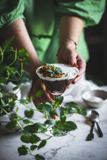 Unrecognizable female putting glass with chocolate and coconut mousse on marble table with green plants - ADSF31513