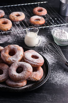 Sweet fried doughnuts served on plate near metal cooling rack and jug of milk on black messy table with powdered sugar - ADSF31540