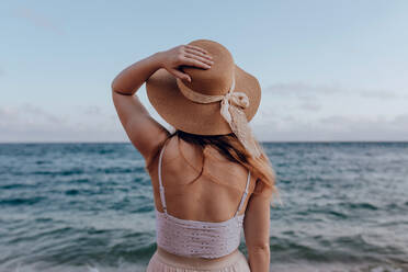 Rückenansicht einer nicht erkennbaren Frau in Sommerkleid und Hut, die am Strand in der Nähe des plätschernden Meeres steht und die malerische Aussicht bewundert - ADSF31563