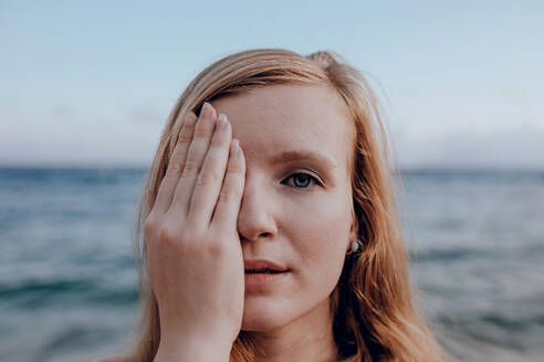 Ruhige junge Frau, die ihr Gesicht mit der Hand bedeckt, während sie in die Kamera schaut, am Meeresufer im Sommerabend auf unscharfem Hintergrund - ADSF31564