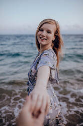 Lächelnde junge Frau in Freizeitkleidung steht am Sandstrand in der Nähe des plätschernden Meeres und hält die Hand einer nicht erkennbaren Person im Sommer - ADSF31565