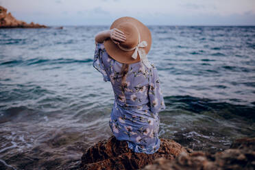 Rückenansicht einer anonymen jungen Frau in Sommerkleid und Hut, die am felsigen Meeresufer sitzt und im Sommerabend in die Ferne schaut - ADSF31567