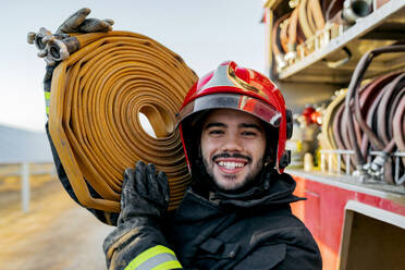 Happy Hispanic firefighter in uniform and helmet carrying hose on shoulder  and gesturing shaka sign while standing near fire engine in daytime stock  photo