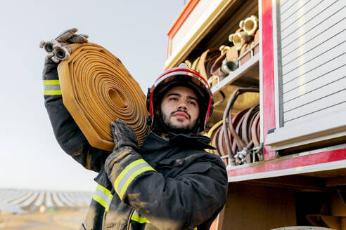 Von unten von einem mutigen männlichen Feuerwehrmann mit Schutzhelm und Uniform, der wegschaut, während er einen großen schweren Schlauch auf der Schulter trägt, in der Nähe eines Feuerwehrautos auf dem Land - ADSF31582