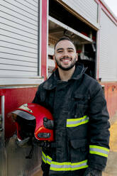 Happy Hispanic firefighter in uniform and helmet carrying hose on shoulder  and gesturing shaka sign while standing near fire engine in daytime stock  photo