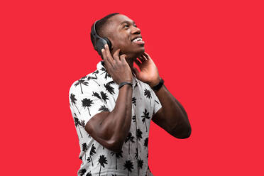 Gut gelaunter afroamerikanischer Mann, der mit kabellosen Kopfhörern genüsslich Lieder hört, während er vor einem roten Hintergrund steht und nach oben schaut - ADSF31599