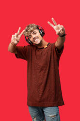 Positiv gestimmter junger Mann mit lockigem Haar, der breit lächelt, während er eine Wiedergabeliste mit drahtlosen Kopfhörern vor rotem Hintergrund abspielt - ADSF31605