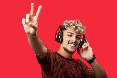 Positiv gestimmter junger Mann mit lockigem Haar, der breit lächelt, während er eine Wiedergabeliste mit drahtlosen Kopfhörern vor rotem Hintergrund abspielt - ADSF31606