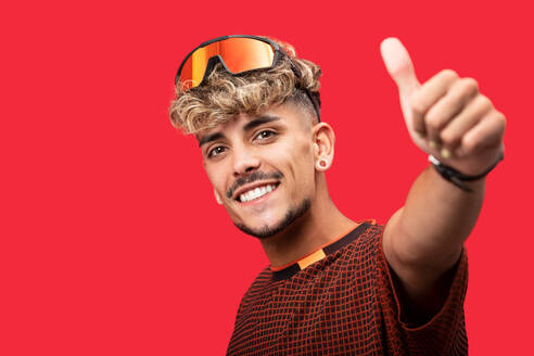 Trendy positive männlich mit lockigem Haar trägt stilvolle T-Shirt und Sonnenbrille zeigt wie Geste vor rotem Hintergrund - ADSF31607