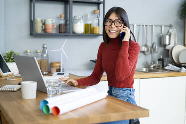 Happy businesswoman with laptop talking on smart phone at home office - GIOF14077