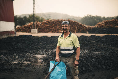 Lächelnder Arbeiter mit Plastiktüte und Werkzeug in einer Kohlefabrik - GRCF01057