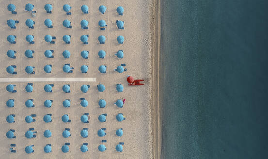Luftaufnahme eines gut sortierten Strandbads mit blauen Sonnenschirmen und Liegestühlen bei Sonnenaufgang an der Küste in Kalabrien, Italien. - AAEF13330