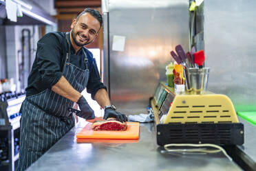 Lächelnder Koch schneidet Fleisch in der Restaurantküche - DLTSF02366