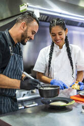Lächelnder Koch und Auszubildender beim Anrichten von Speisen in einer Großküche - DLTSF02381