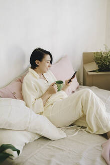 Mature woman with coffee cup using smart phone in bedroom - SEAF00011