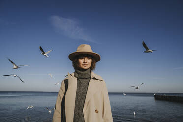 Woman in overcoat and hat standing in front of seagulls flying over sea - SSGF00246