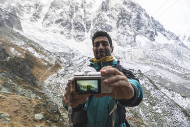 Smiling man taking selfie through camera in front of snowcapped mountain, Chamonix, France - JAQF00883