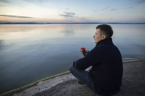 Man drinking tea sitting on rock by lake at sunset - LLUF00316
