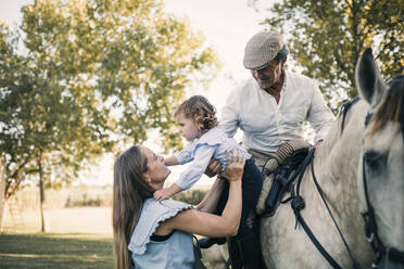 Mutter trägt Tochter, während älterer Mann auf Pferd sitzt - GRCF01082