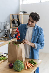 Young man removing cherry tomatoes from bag at kitchen counter - GIOF14110