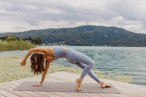 Active woman practicing yoga on jetty by lake - DAWF02180