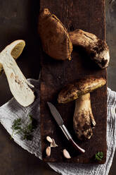 Draufsicht auf roh geschnittene Steinpilze (Boletus edulis) auf einem hölzernen Schneidebrett mit Knoblauch und Petersilie in einer hellen Küche während des Kochvorgangs - ADSF31647