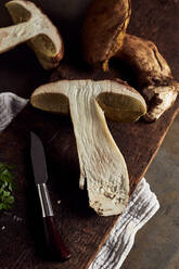 Top view of raw cut Boletus edulis mushrooms on wooden chopping board with garlic and parsley in light kitchen during cooking process - ADSF31648