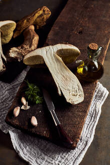 Top view of raw cut Boletus edulis mushrooms on wooden chopping board with garlic and parsley in light kitchen during cooking process - ADSF31650