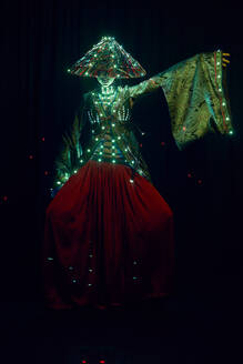 Anonyme Frau in authentischem Outfit und vietnamesischem Hut mit leuchtender Beleuchtung in einem dunklen, modernen Studio auf schwarzem Hintergrund während einer Performance - ADSF31681