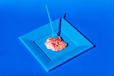 Haufen von rosa rohen Gehirnen auf blauem Teller mit Plastikgabel auf blauem Hintergrund in hellen modernen kreativen Studio serviert - ADSF31685