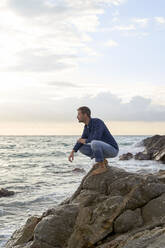 Man crouching on rock by sea at beach - VEGF05183