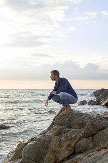 Mann hockt am Strand auf einem Felsen am Meer - VEGF05183