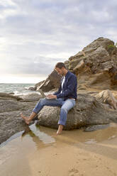 Man sitting on rock using laptop at beach - VEGF05195