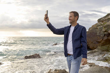 Lächelnder Mann nimmt Selfie durch Smartphone am Strand - VEGF05196