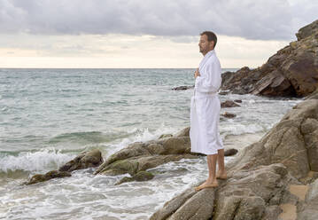 Mann im Bademantel steht auf einem Felsen am Strand - VEGF05199