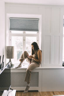 Frau mit Smartphone auf der Fensterbank zu Hause sitzend - DAWF02202