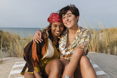 Lächelndes lesbisches Paar umarmt sich gegenseitig am Strand sitzend - JRVF02041