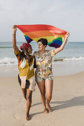 Carefree lesbian couple with rainbow flag walking at beach - JRVF02061