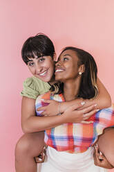 Woman piggybacking lesbian friend against pink background - JRVF02082