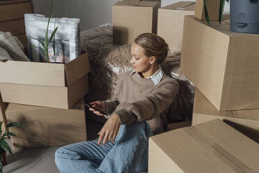 Young woman using smart phone amidst cartons in new home - VPIF05187