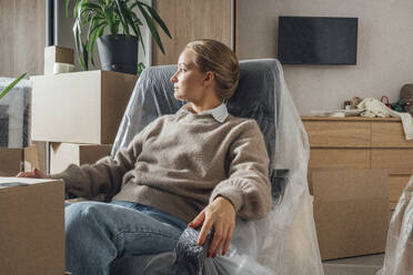 Nachdenkliche Frau ruht sich auf einem in Plastik eingewickelten Sessel in ihrem neuen Zuhause aus - VPIF05200