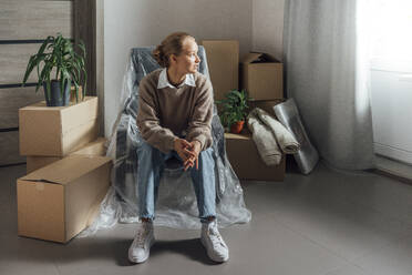 Junge Frau sitzt auf einem in Plastik eingewickelten Sessel in einer umgesiedelten Wohnung - VPIF05207